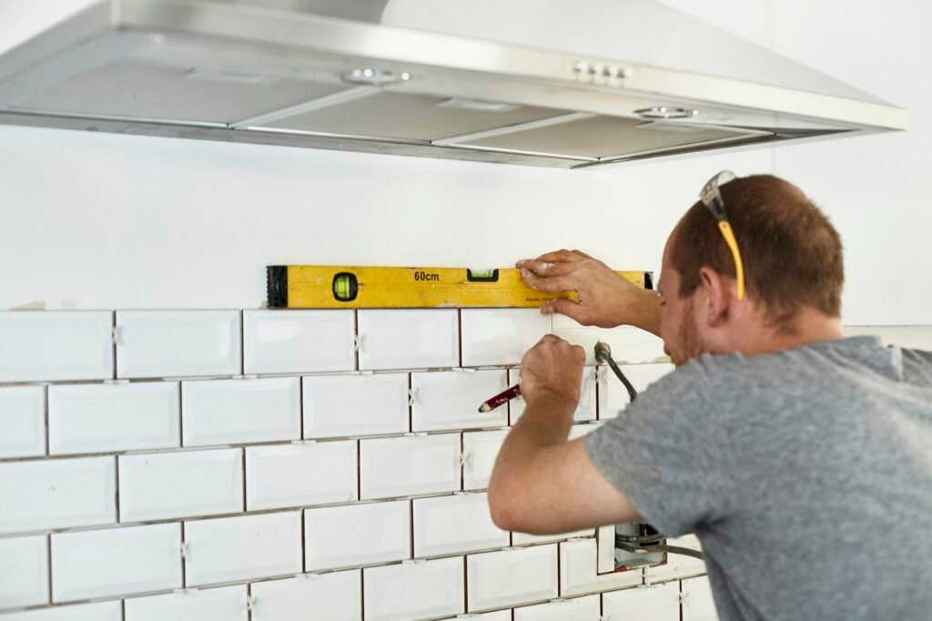 A man working in a new kitchen, a tiler applying tiles to the wall behind the cooker.