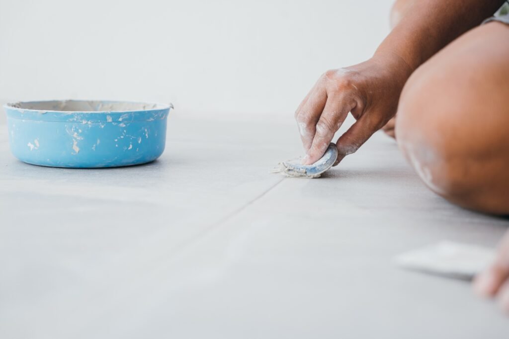 A hand with a rubber trowel smoothing mortar between gray ceramic floor tiles